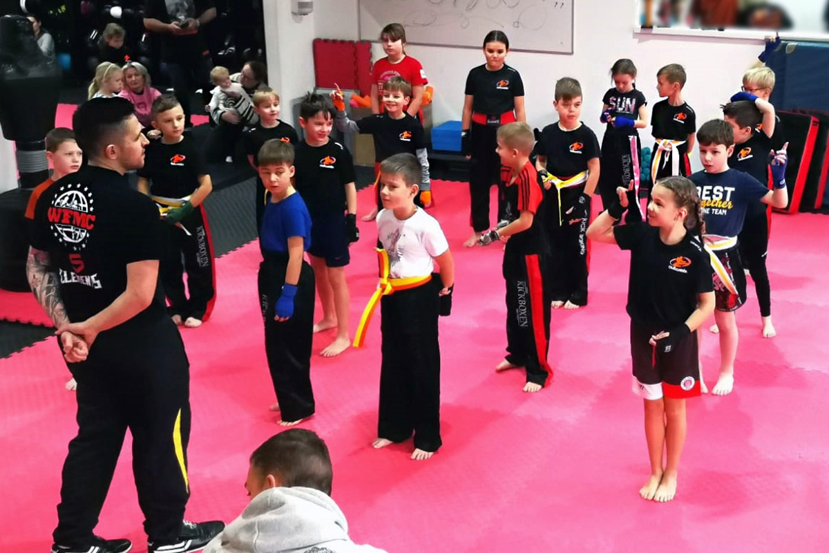 Startseite 5 Mehrere Kinder stehen in Sportkleidung mit Gürteln auf einer Matte und hören einer erwachsenen Person zu.